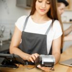 Coffee Business Concept - Beautiful female barista giving payment service for customer with credit card and smiling while working at the bar counter in modern coffee shop.