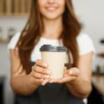Coffee Business Concept - Beautiful Caucasian lady smiling at camera offers disposable take away hot coffee at the modern coffee shop